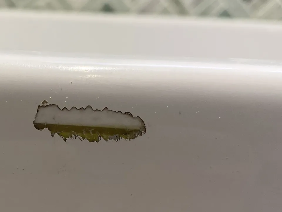 Side of a white porcelain bath below a green herringbone tile wall, with a long chip showing the dark base underneath