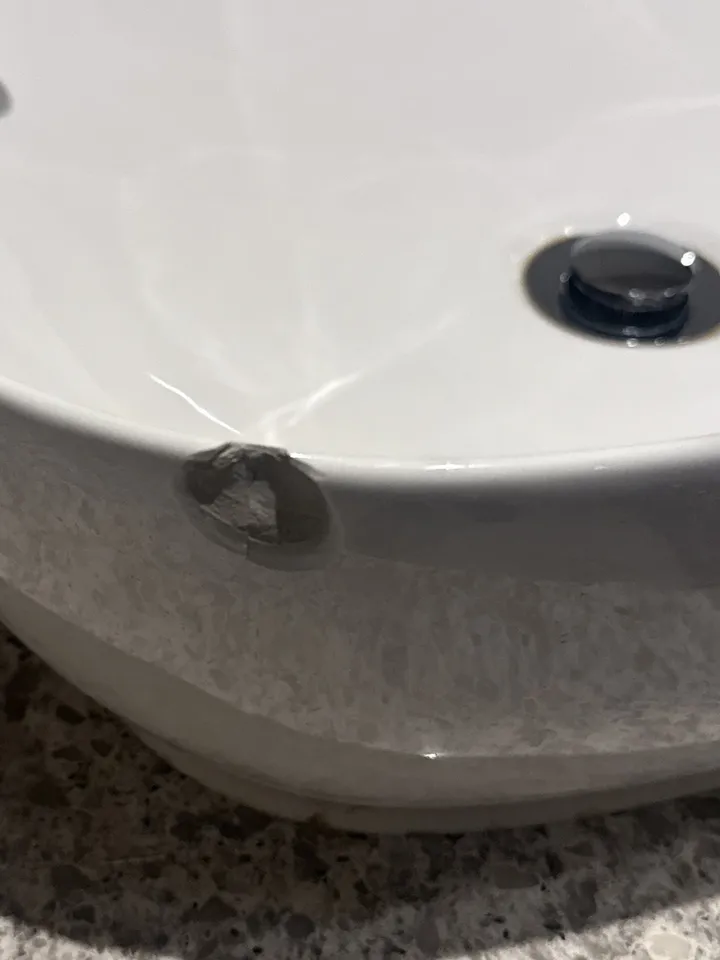 White porcelain vessel basin on a speckled vanity top with a chip out of the side, showing grey body underneath